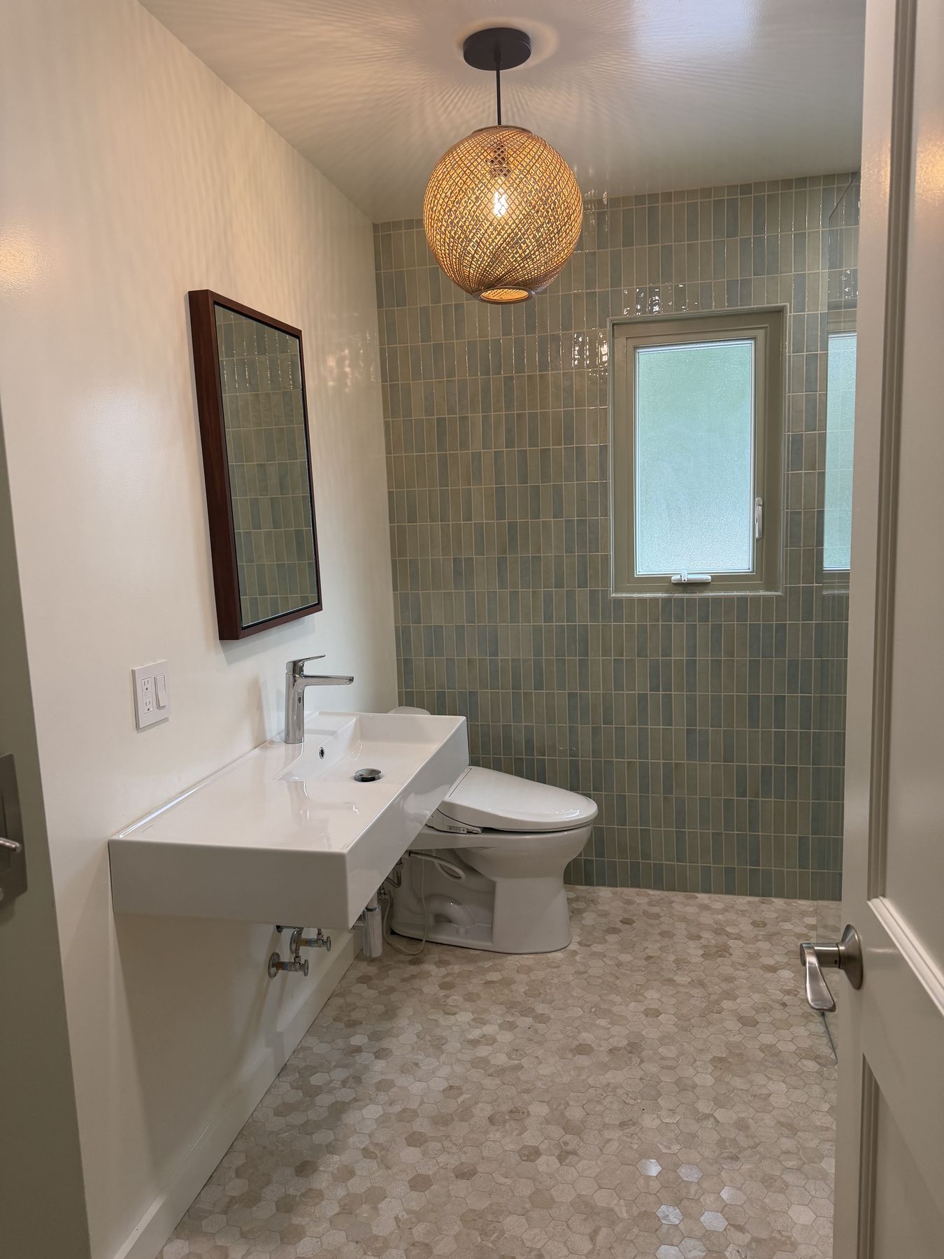 Remodeled bathroom with green vertical tile, rattan pendant, and stone hex floor